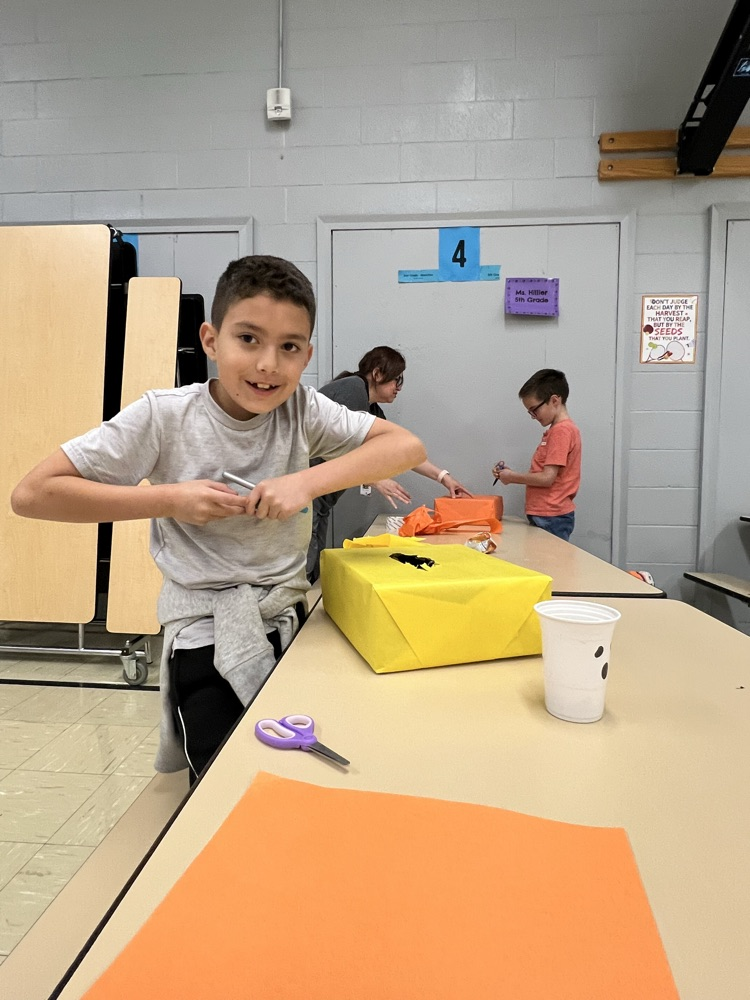 A boy holds scissors while another holds a paper bag at a table in a classroom.