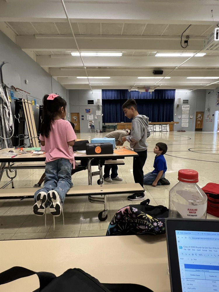 Students work in a gym with a blue curtain at the back. Two people sit on a bench, and one stands at a table.