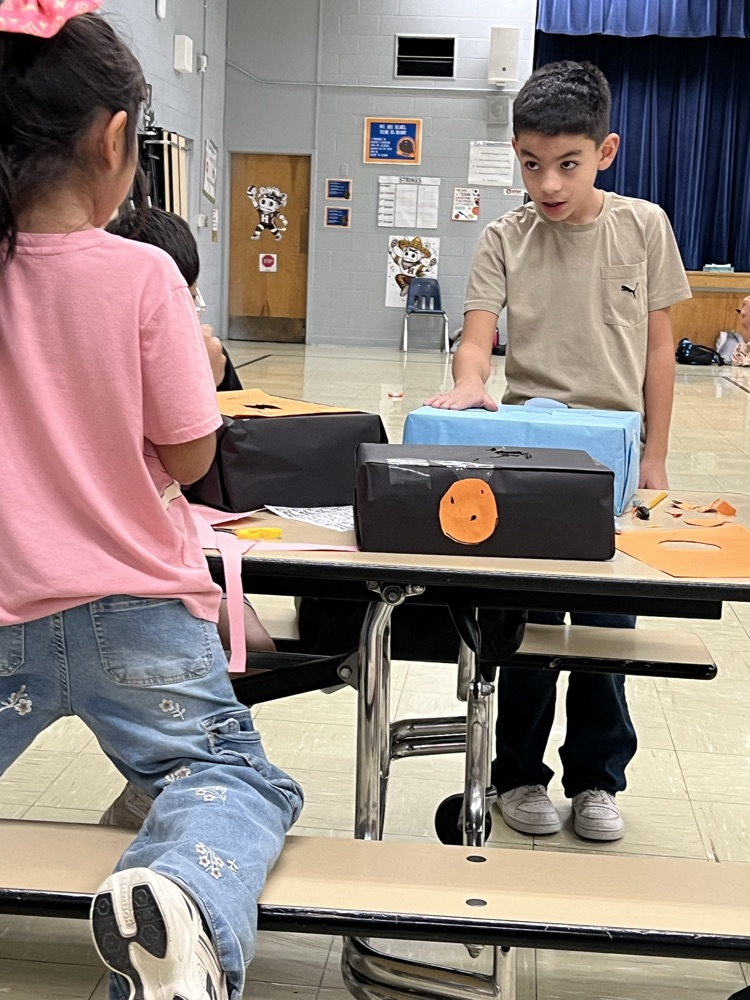 Two students, one in pink, the other in brown, standing at a table with two boxes.