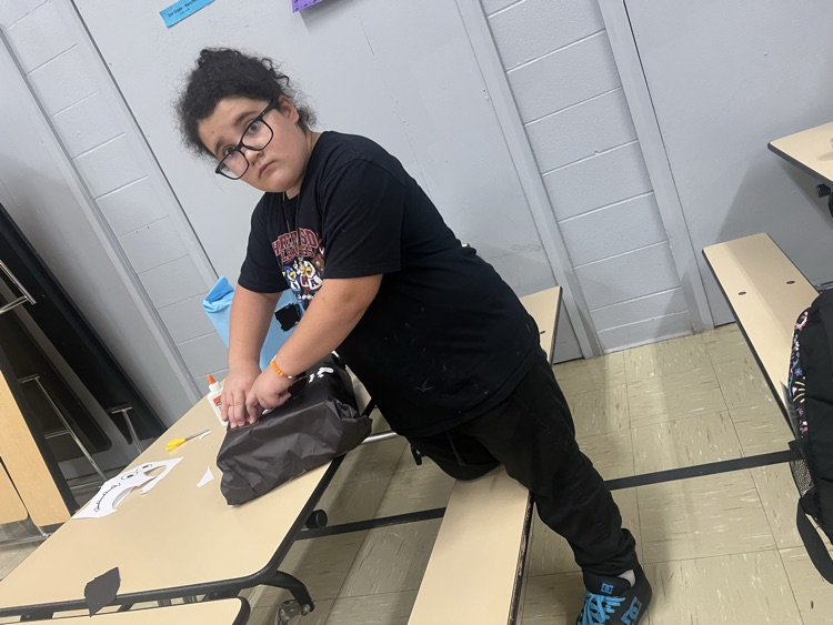 Person wearing glasses and a black shirt stands at a table, adjusting a backpack. Behind them, benches and a white wall.
