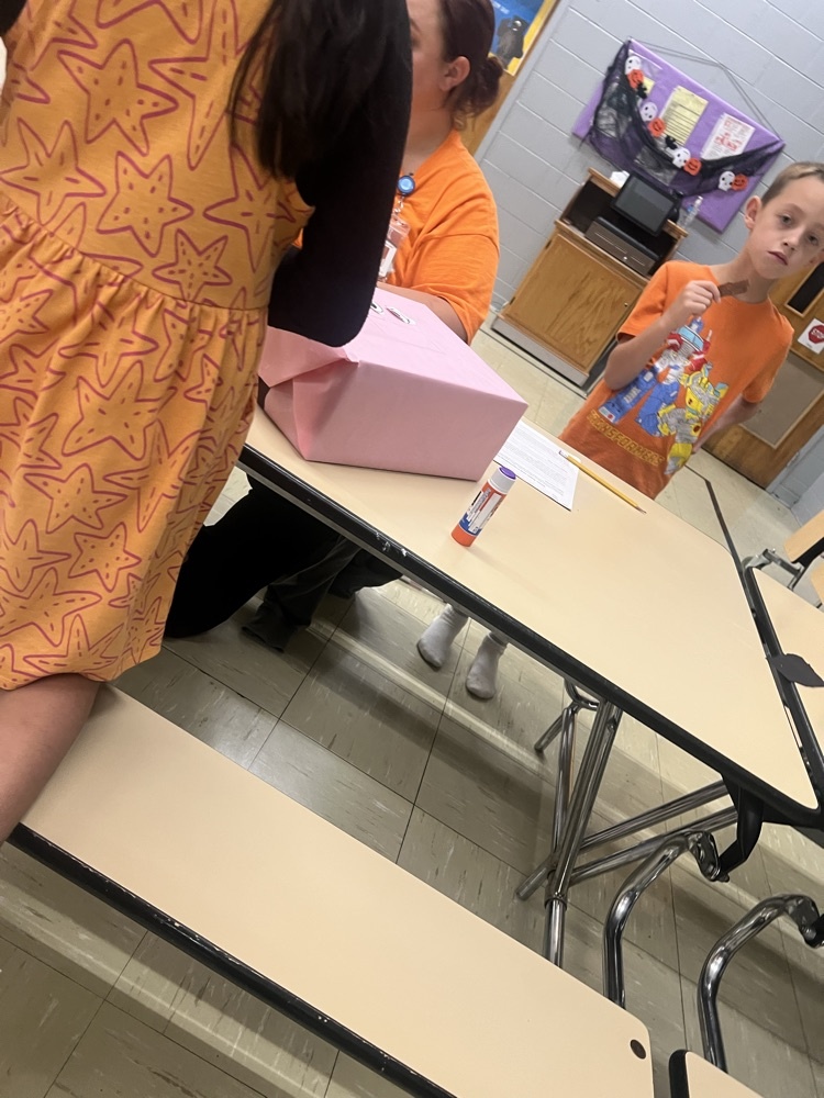 Two students in a classroom, one with a box on a table, another holding a marker.