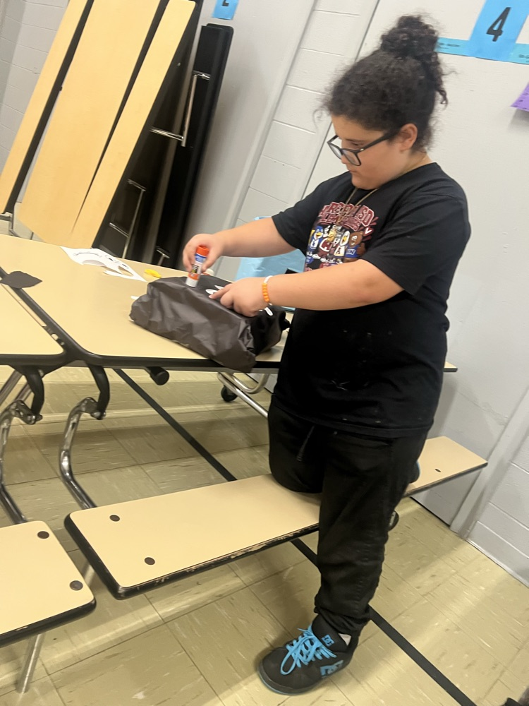 A person in a classroom sits on a bench and holds a bag, possibly repairing it.