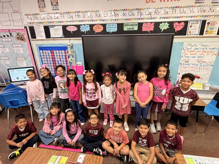 Classroom with a large screen, children in pink and maroon shirts, and chairs and desks.