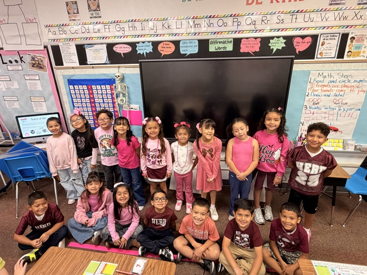 A classroom with children seated and standing, some wearing pink shirts. A large screen is on the wall, and a teacher is present.
