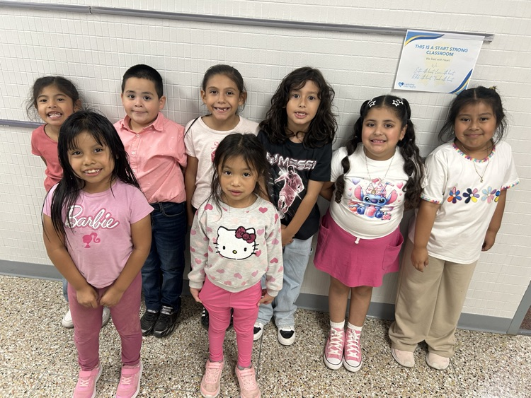 Eight young girls in different colored tops and pants stand next to each other in front of a wall.