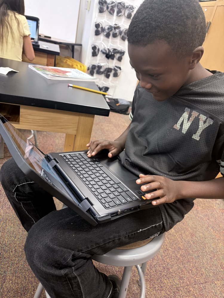 Child sits on stool at table, focused on laptop, wearing a gray shirt with the letters NY.