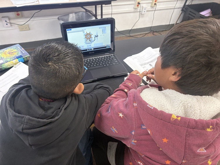 Two children are sitting in chairs in front of a laptop on a desk. One child wears a sweatshirt.