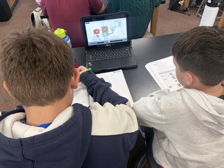 Two boys seated at a table, one looking at a laptop screen displaying a diagram.