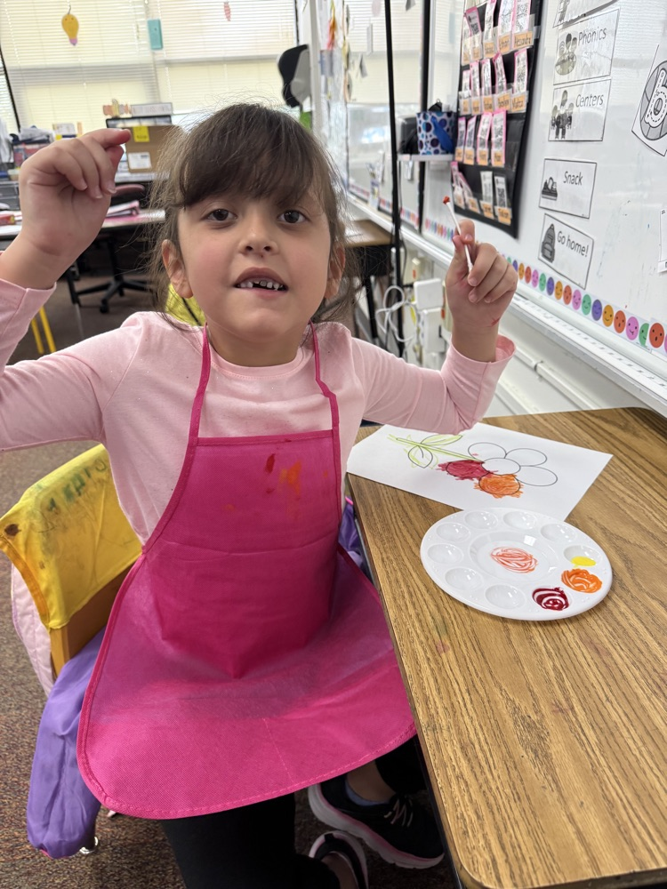 Girl in pink apron sits at a table holding a paintbrush. A palette with paint sits in front of her.