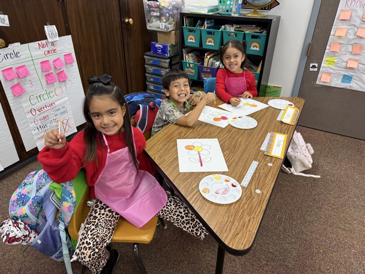 Three children seated at a table with a backpack, books, and paper. One holds a paintbrush.