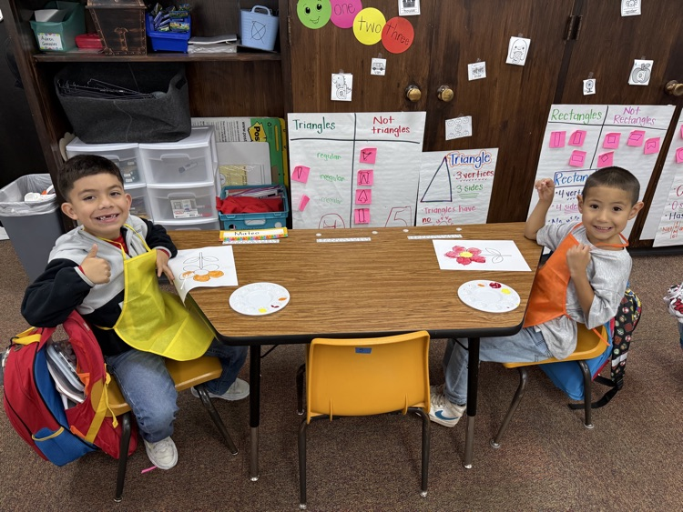 Two children at a desk, one holding a book, and both smiling. A backpack and book bag sit behind them.