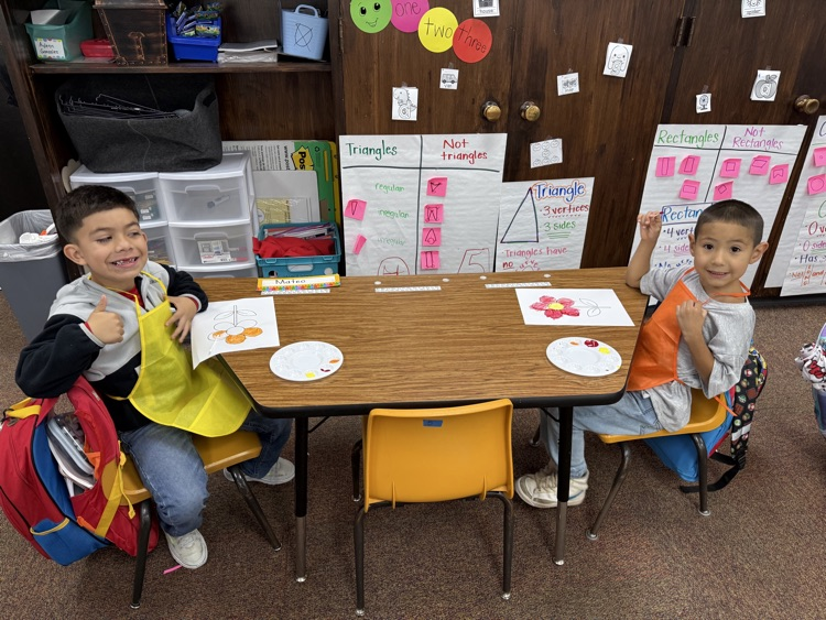 Two young boys sitting at a small table in a classroom, smiling and holding papers.