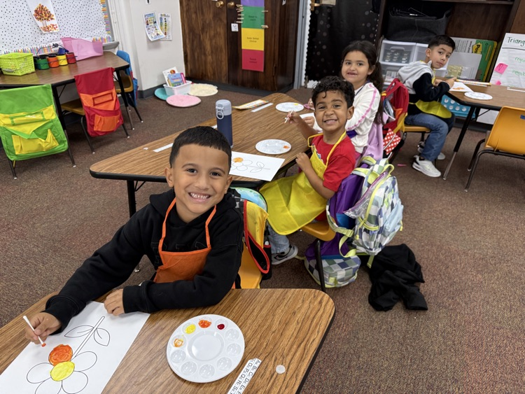 Several children sit at tables in a classroom. One child is painting on paper with a palette.