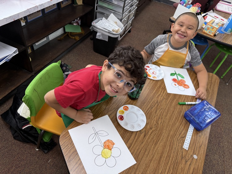 Two boys sit at a table with a drawing of a flower, paint, and a paintbrush.