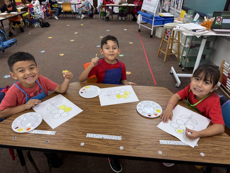 Three young boys seated at a wooden table with paint palettes and paper, working on art projects.