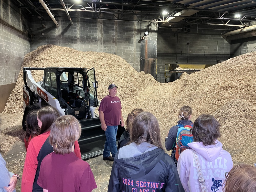 Honors Program students and heating plant worker by heavy machinery and wood chips