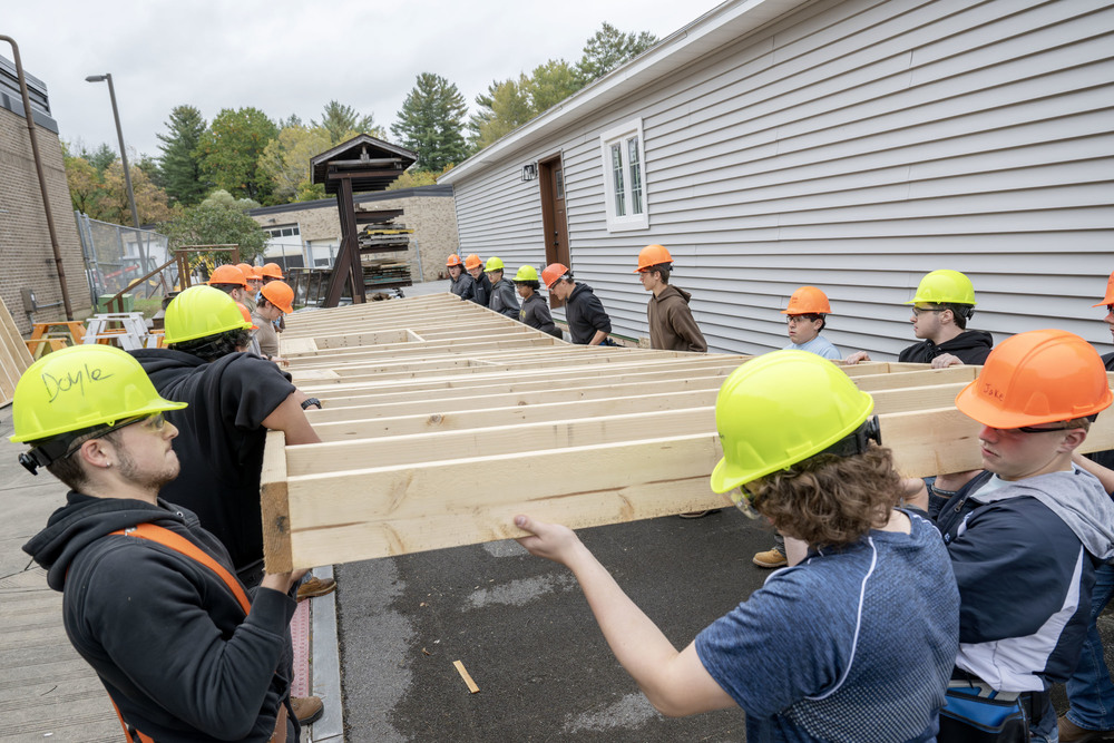 Many students in hardhats are carrying a large piece of a house they are building