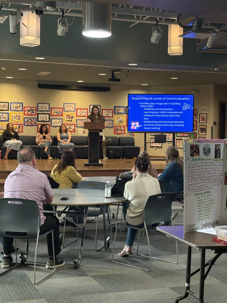 Several people sitting at tables, a few people sitting on stage and one person speaking at a podium on stage