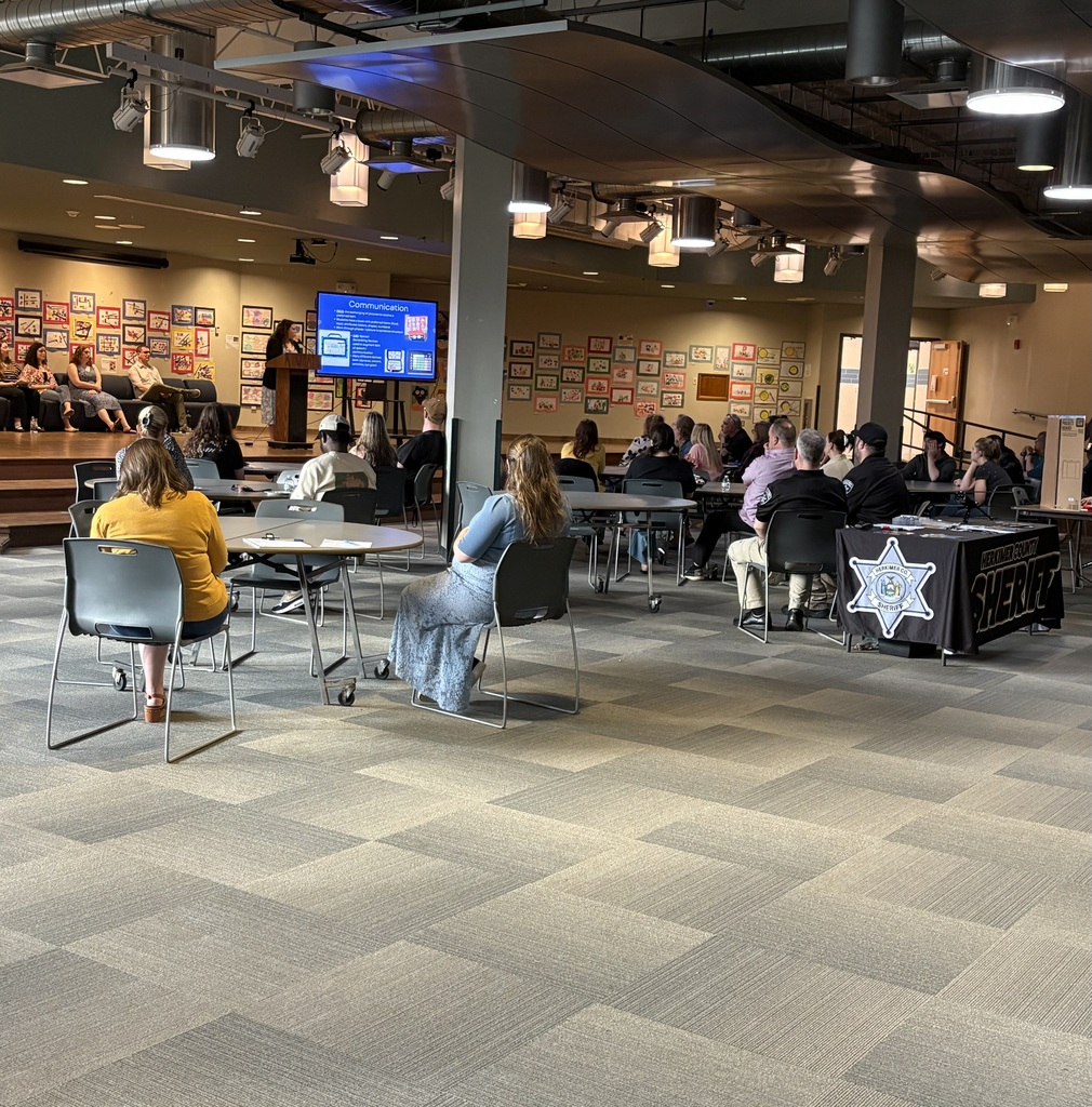 WEB Complex lobby with people sitting at tables and on stage with a person speaking at podium on stage