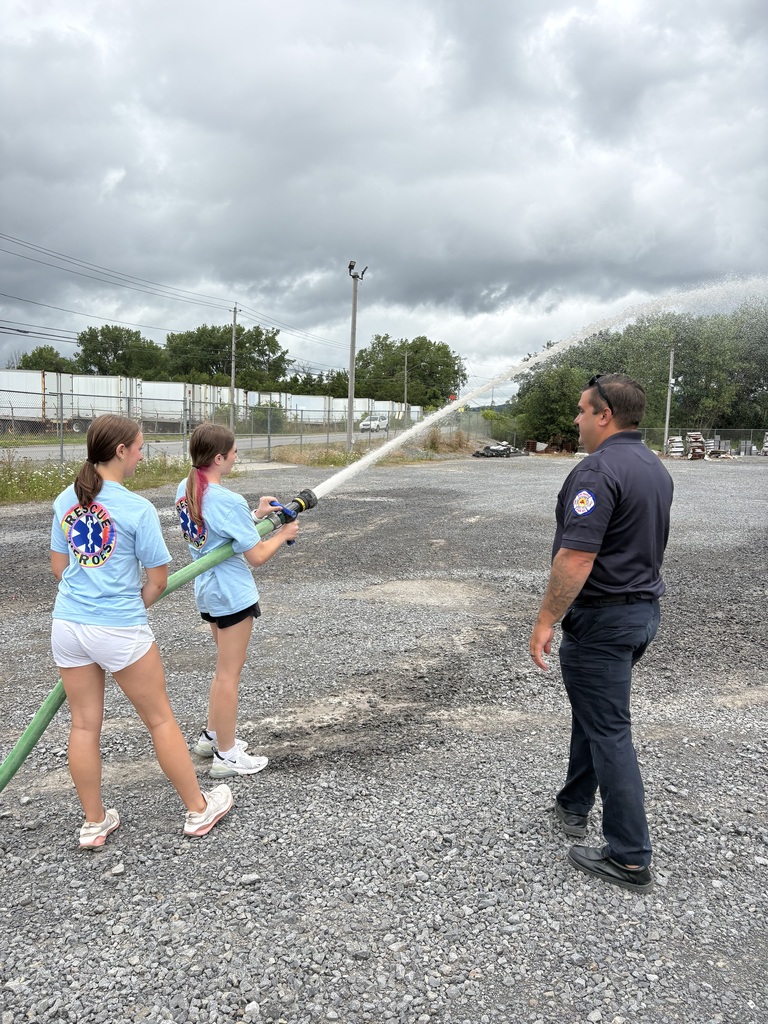 Two students spraying a hose with a firefighter nearby
