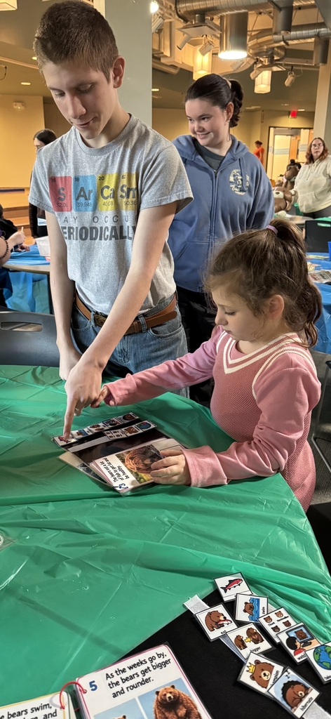 Person pointing at a bear book to a younger student, and another student in the background