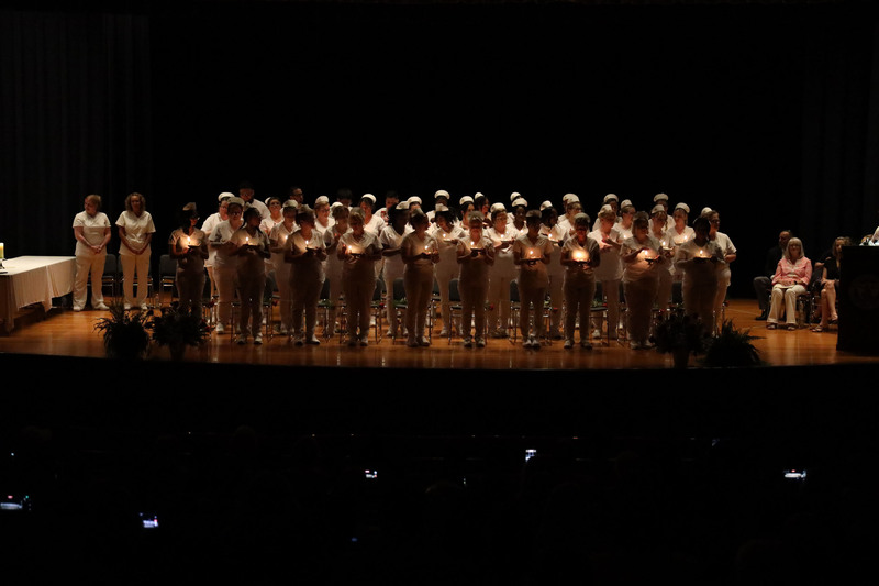 Adult LPN graduates standing on stage holding candles in a darkened auditorium for a candle-lighting ceremony