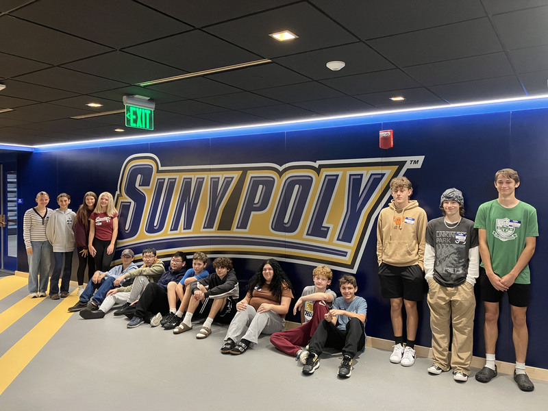 15 students standing by wall decoreated with the words "SUNY Poly"