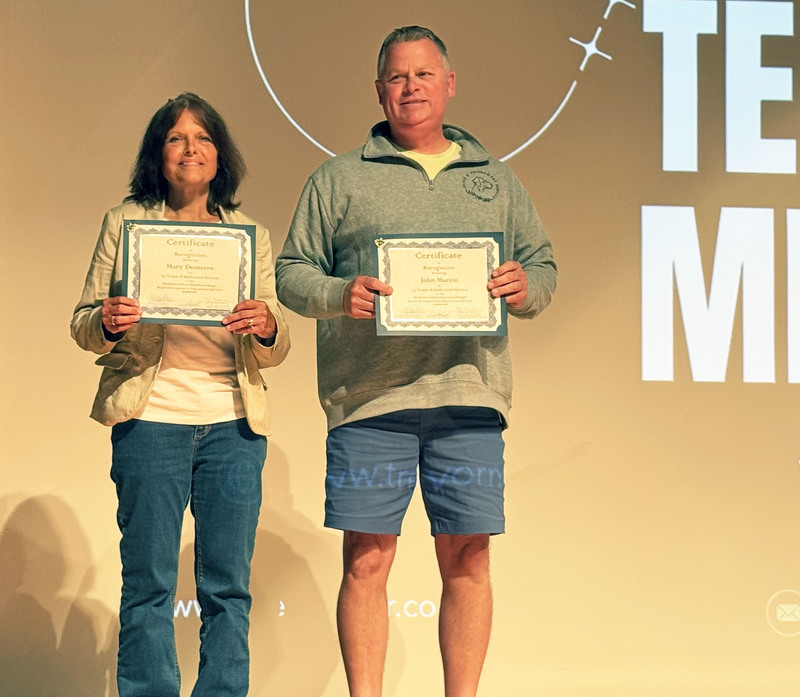 Mary Demere and John Martin on stage together holding certificates for years of service