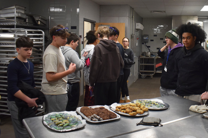 Two students presenting in culinary kitchen to others looking on with cookie platters nearby