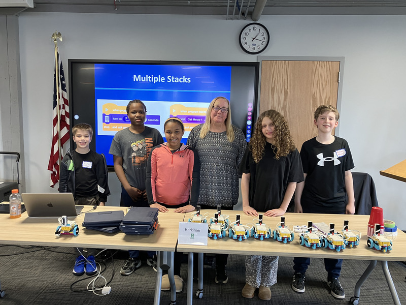 Students and teacher and presenter posing by table with robots and computers