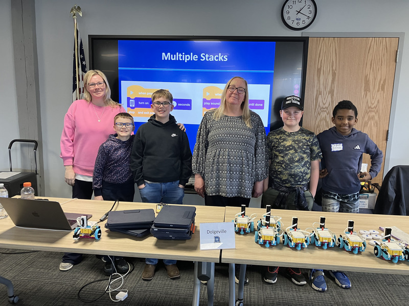 Students and teacher and presenter posing by table with robots and computers