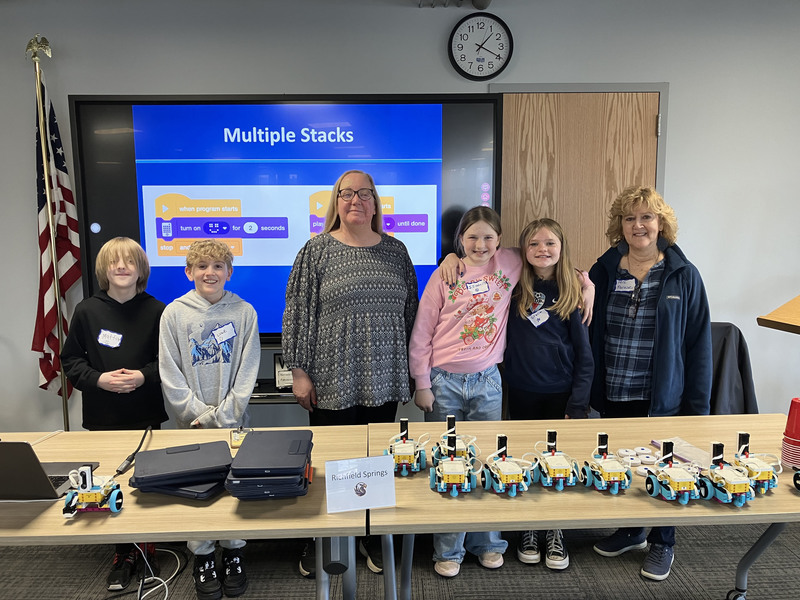 Students and teacher and presenter posing by table with robots and computers