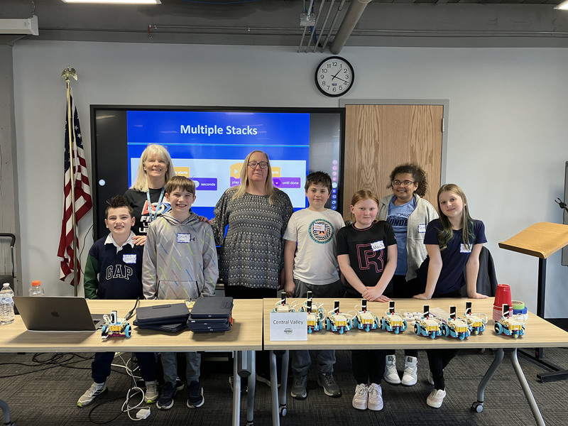 Students and teacher and presenter posing by table with robots and computers