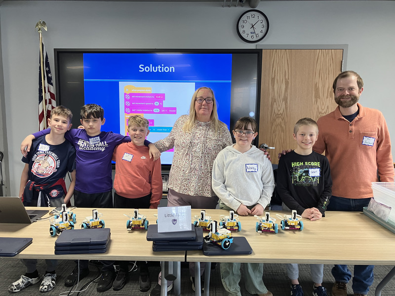 Students and teacher and presenter posing by table with robots and computers