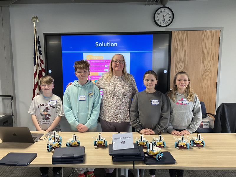 Students and teacher and presenter posing by table with robots and computers