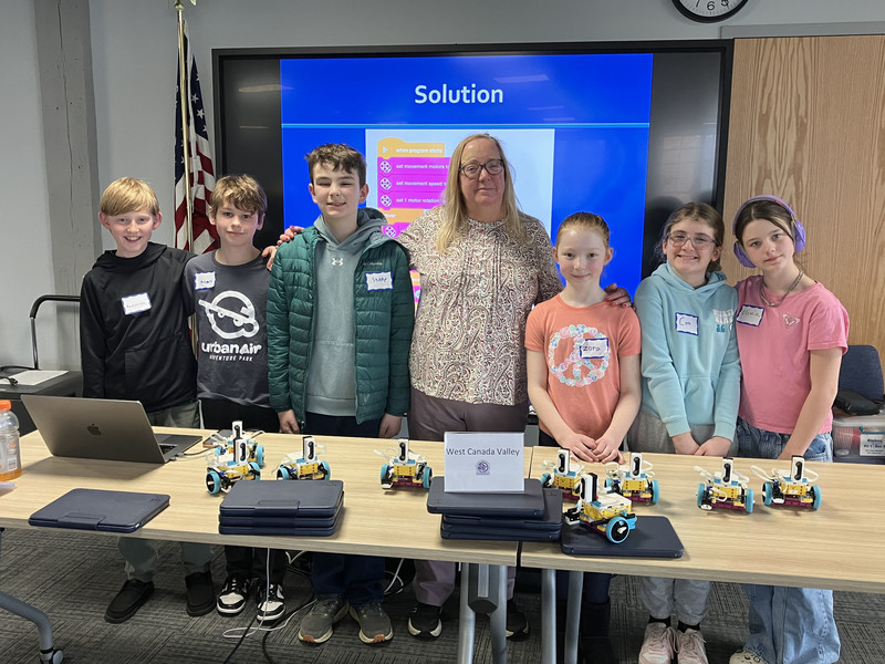 Students and teacher and presenter posing by table with robots and computers
