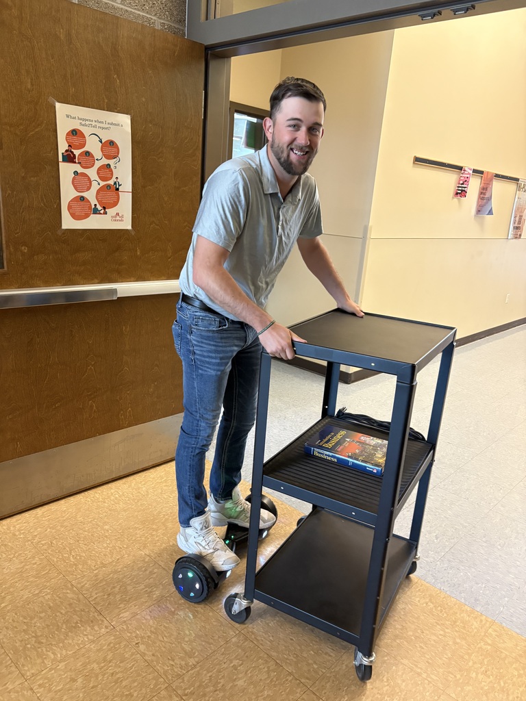 Mr. Marzolf on a hoverboard pushing a cart with a book