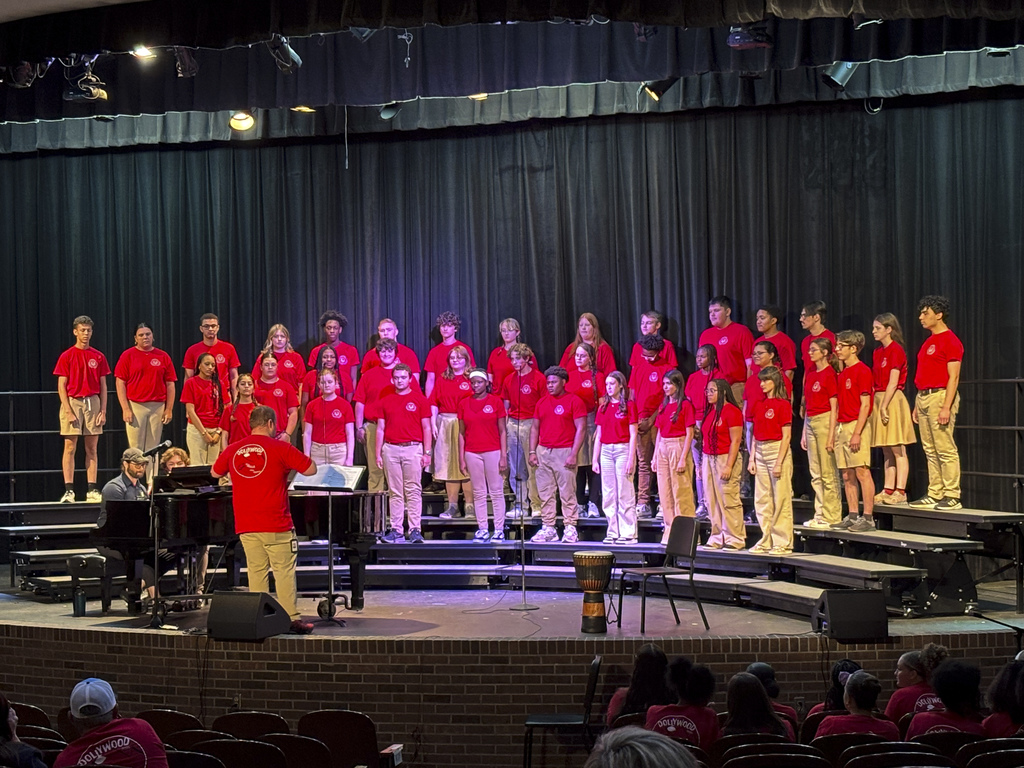 A choir wearing red shirts and khaki shorts performs on stage with a pianist and drum.