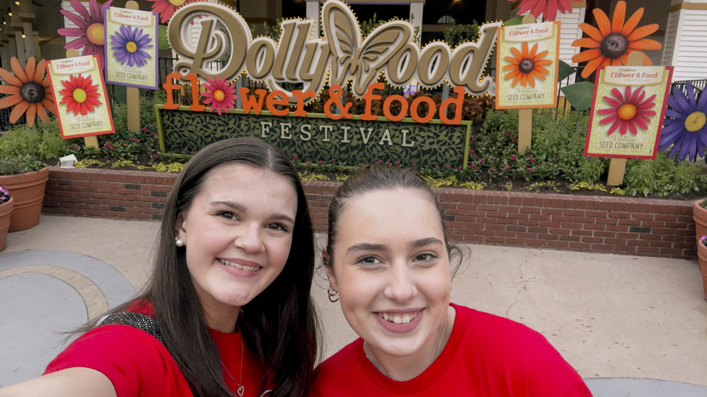 Two women in red shirts smile for a selfie at the DollyWood Flower & Food Festival, with colorful flowers and signs in the background.