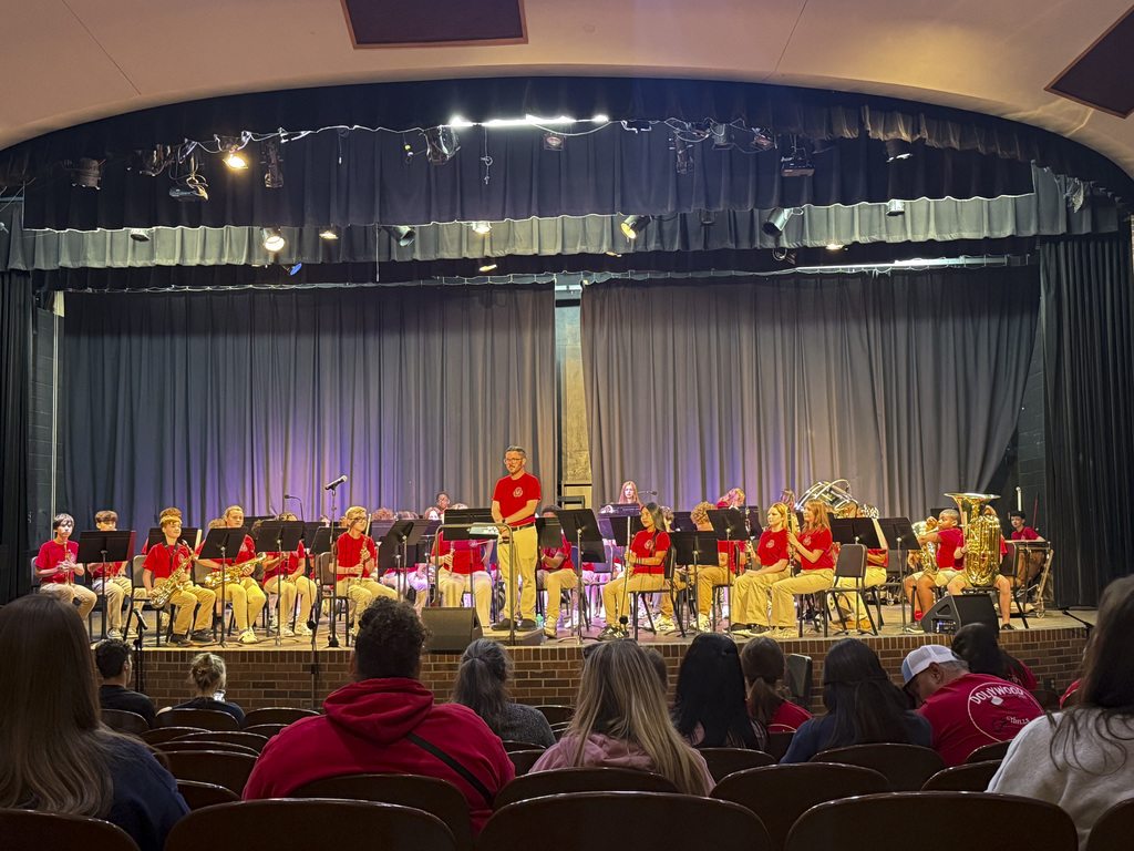 A band performs on stage with red shirts and yellow pants, under purple stage lights. An audience watches from chairs.