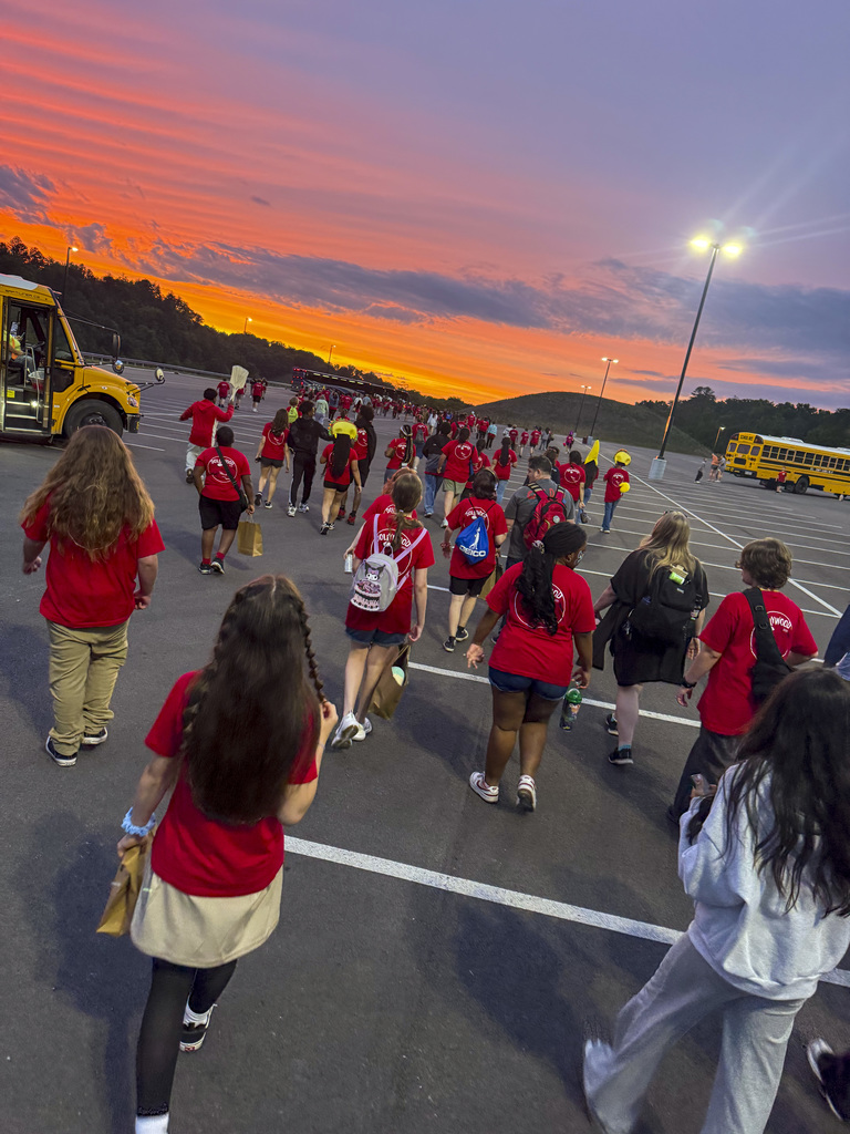 People in red shirts walk in a parking lot at sunset, with two yellow school buses parked.