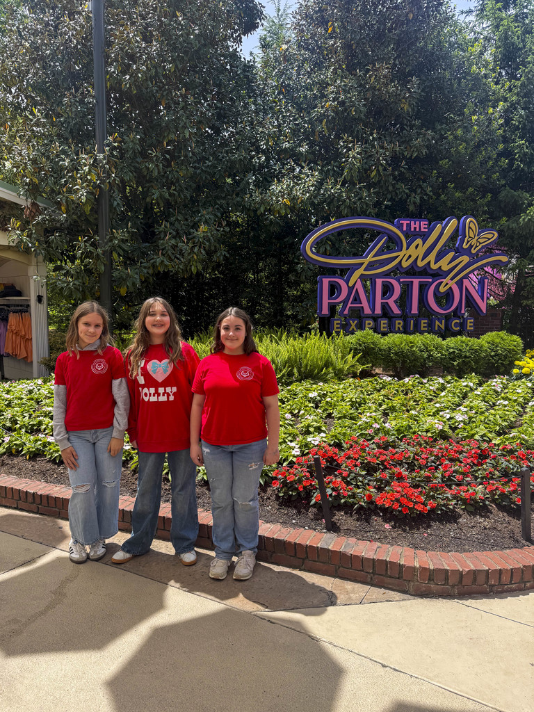 Three girls in matching red shirts stand before a garden with red and white flowers.