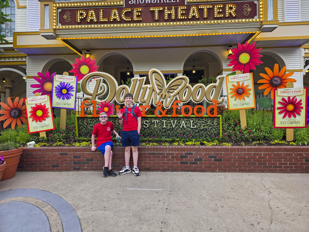 Two children stand in front of a flower-decorated sign with "DollyWood Food & Food Festival" and a building with "Palace Theater" above it.