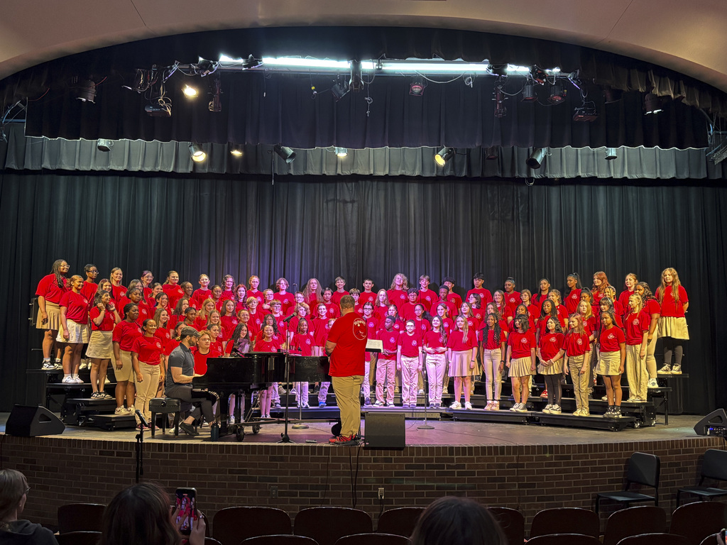 A choir in red and white performs on a stage with a pianist in front of a seated audience.