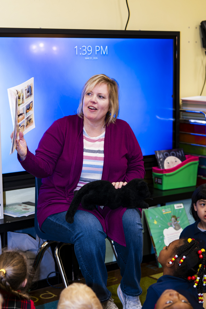 Woman in purple cardigan sits in a chair and reads a book to children in a classroom.