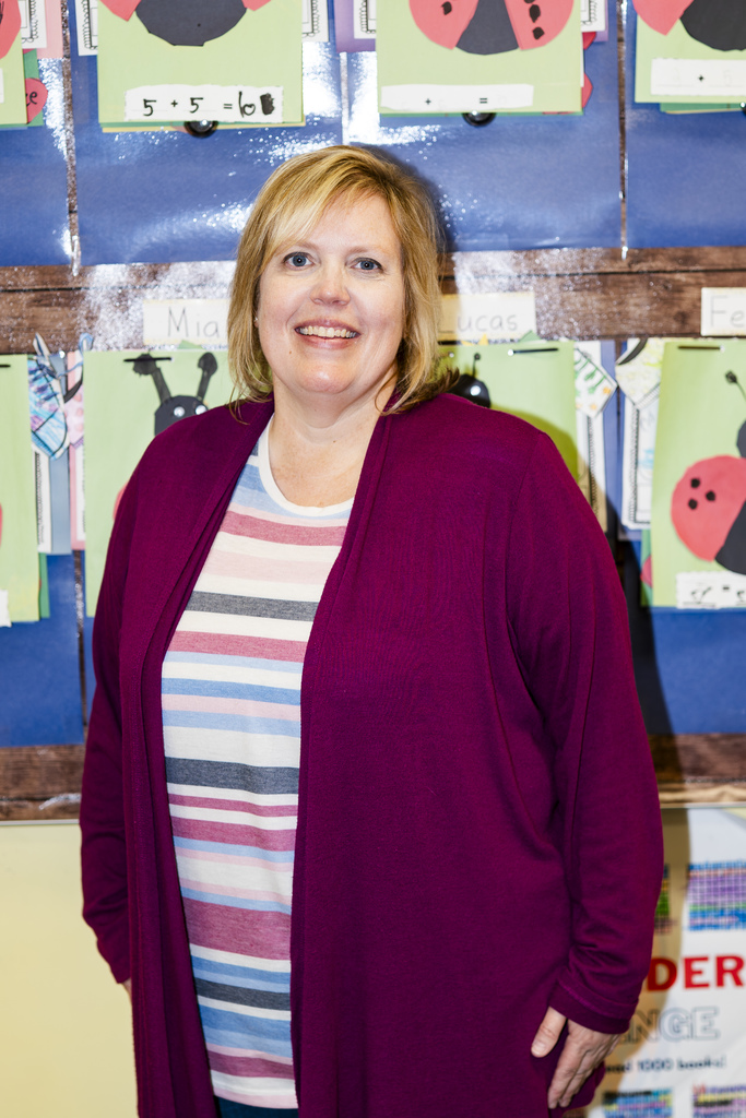 A woman wearing a purple cardigan and striped shirt smiles while standing in front of a bulletin board with colorful children's artwork.