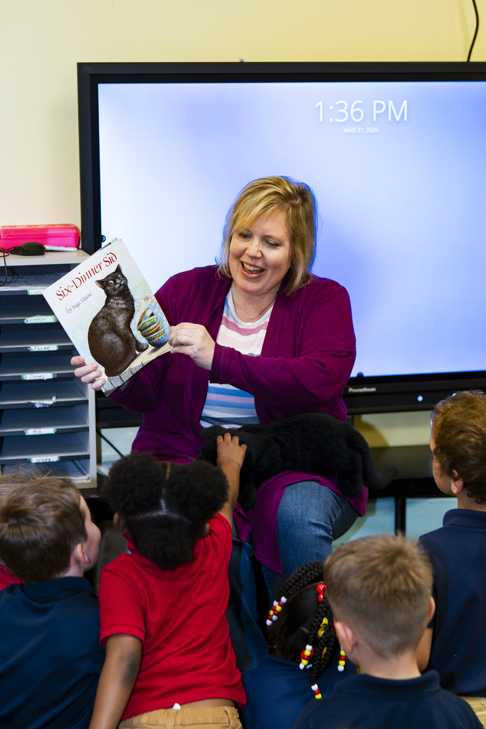 A woman sits with children, reading a book with a cat. They sit in a classroom.