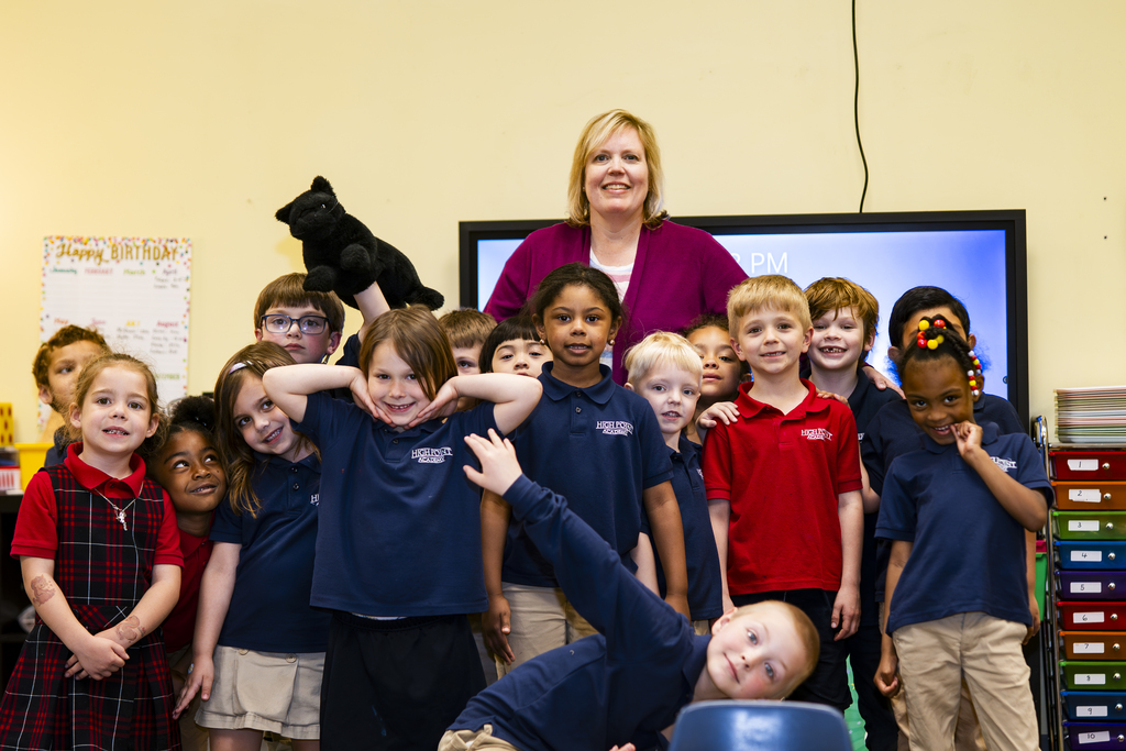 A group of children in a classroom, with a woman standing behind them. The children are smiling and posing for a photo.
