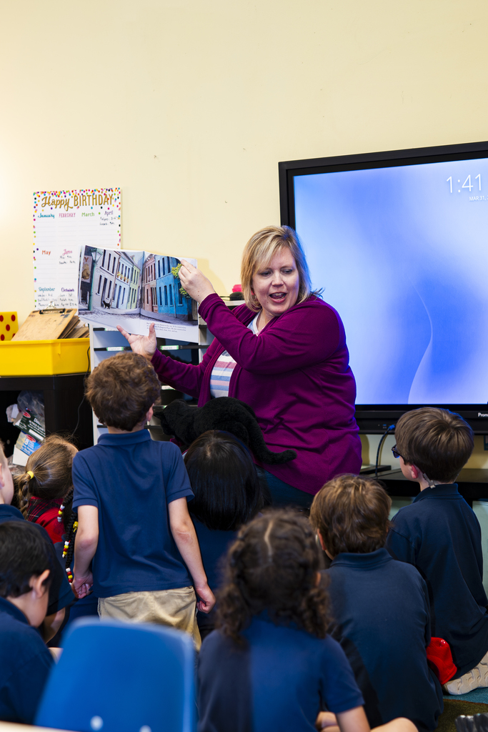 Woman stands with an open book while a group of children listen and watch a large screen.