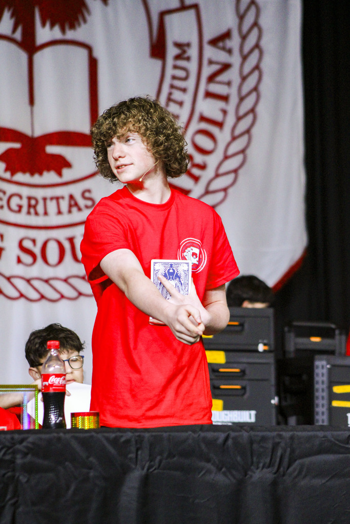 A person stands at a table with cards, wearing a red shirt. A Coca-Cola bottle is on the table.